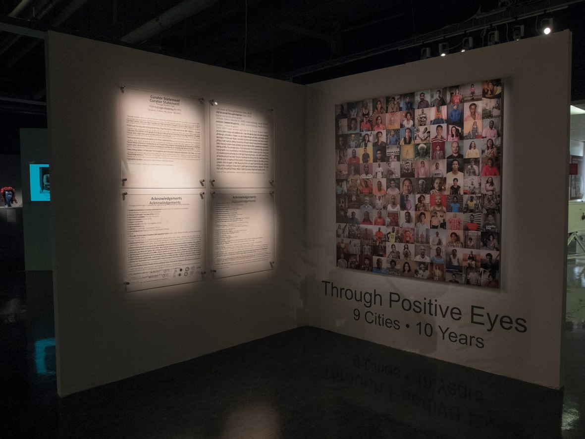 Introductory text on the left wall and Gideon Mendel’s composite portraits above the *Through Positive Eyes* title on the right